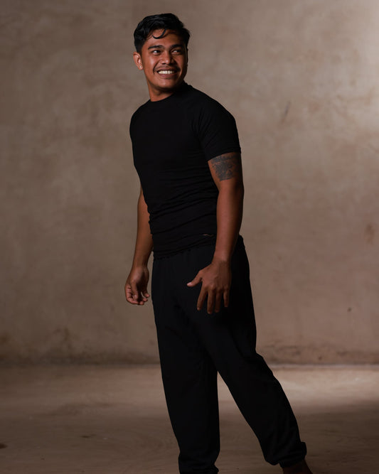 Man wearing a black t-shirt and black pants standing against a beige wall.